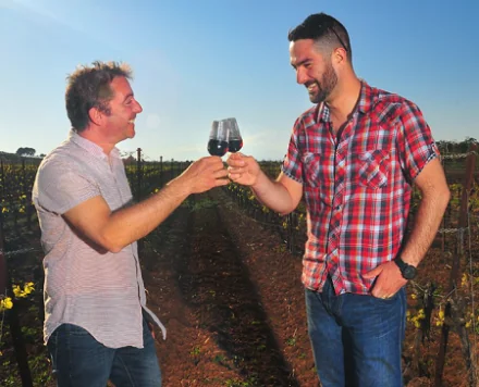 Les deux vignerons (Francois Delhon et Jean-Philippe Leca) trinquent avec un verre de vin rouge, debout au milieu des rangs de vigne au soleil. Photo authentique, chaleureuse, qui incarne l'equipe et la convivialite du domaine. Seul vrai portrait humain disponible sur le site - tres valorisant.