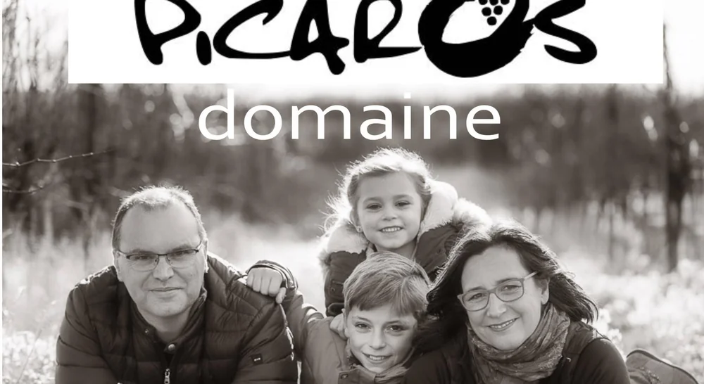 Portrait de famille en noir et blanc dans les vignes : Pierre-Yves Rouille et Caroline Vioche (les vignerons) entoures de leurs deux enfants, tous souriants, allonges dans une parcelle hivernale. Logo PICARO'S et mot 'domaine' surimprimes en haut. Version la plus large et la mieux definie de la phot