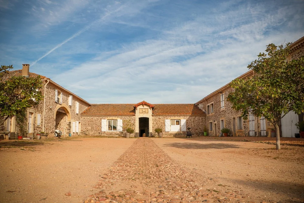 Vue frontale du mas en pierre : cour pavée symétrique, bâtiments anciens avec porte centrale et fronton, ciel bleu, arbres en cadre. Hero shot du domaine, photo professionnelle (Maryne Torchy).