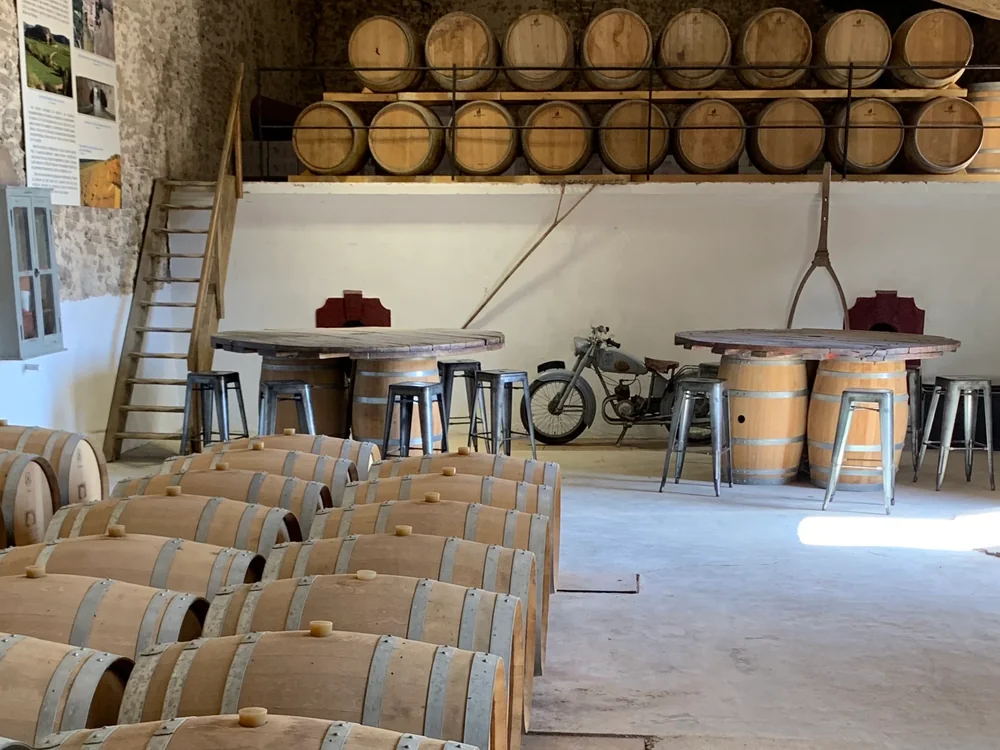 Chai/caveau de dégustation : rangées de barriques de chêne au sol et sur racks, tables-tonneaux avec tabourets métalliques, vieille moto en décor, panneaux d'interprétation au mur. Authentique, légèrement amateur mais représentatif du lieu de dégustation.