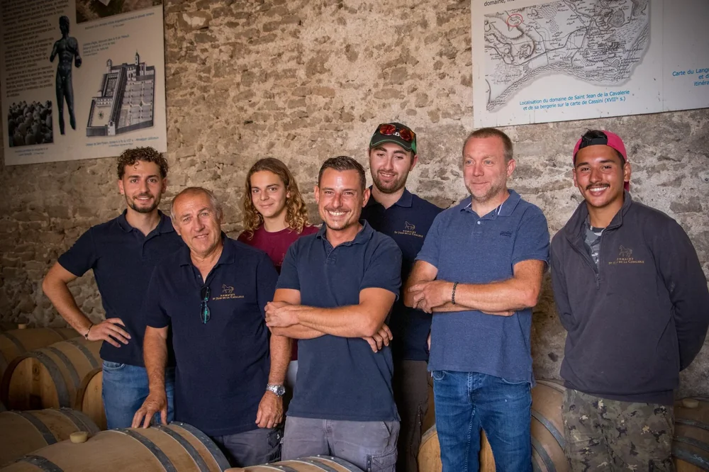 Photo de groupe de l'équipe/famille du domaine (7 personnes, plusieurs en polos brodés au logo cheval) dans le chai, debout au milieu des barriques de chêne, devant des panneaux d'interprétation historique (dont la carte de Cassini du domaine). Image très valorisante : l'humain et le terroir réunis.
