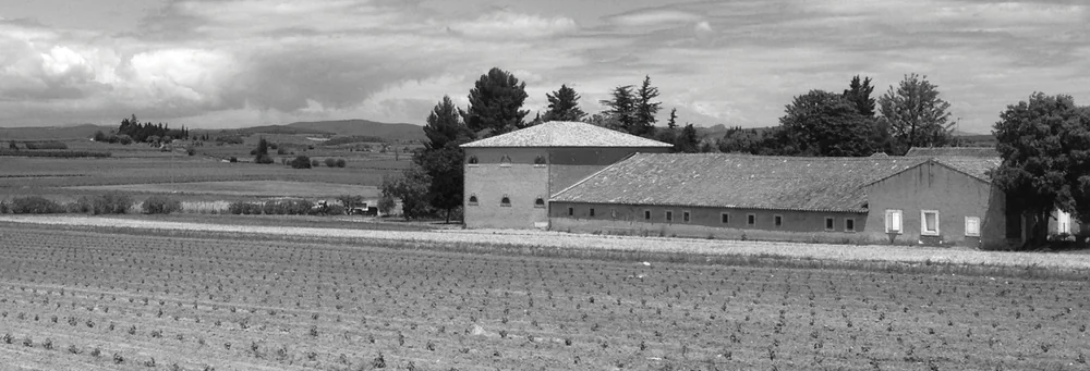Vue d'ensemble noir et blanc du mas/château du domaine : grand bâtiment viticole en pierre à toiture pyramidale, dépendances, entouré de vignes labourées et de cyprès, collines au loin. Belle vue architecturale d'ensemble du domaine.