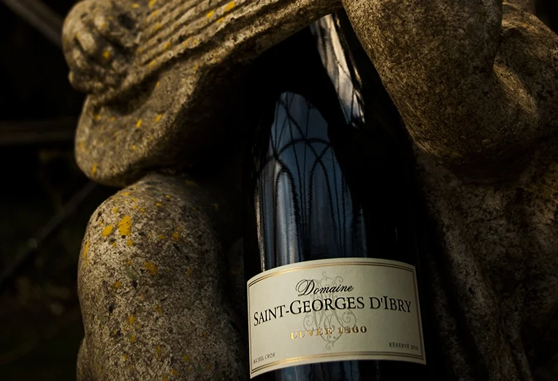 Belle photo en situation : bouteille de Cuvée 1860 (rouge réserve) blottie contre une statue de pierre couverte de lichen, éclairage clair-obscur. Packshot lifestyle élégant et atmosphérique, au-delà de la simple étiquette.