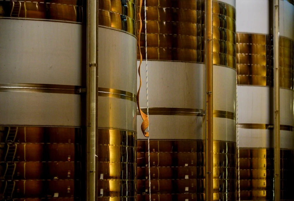 Chai : rangée de cuves de vinification en inox brillantes reflétant des tons dorés et cuivrés, avec une tireuse à échantillon. Image graphique et qualitative de l'outil de production.