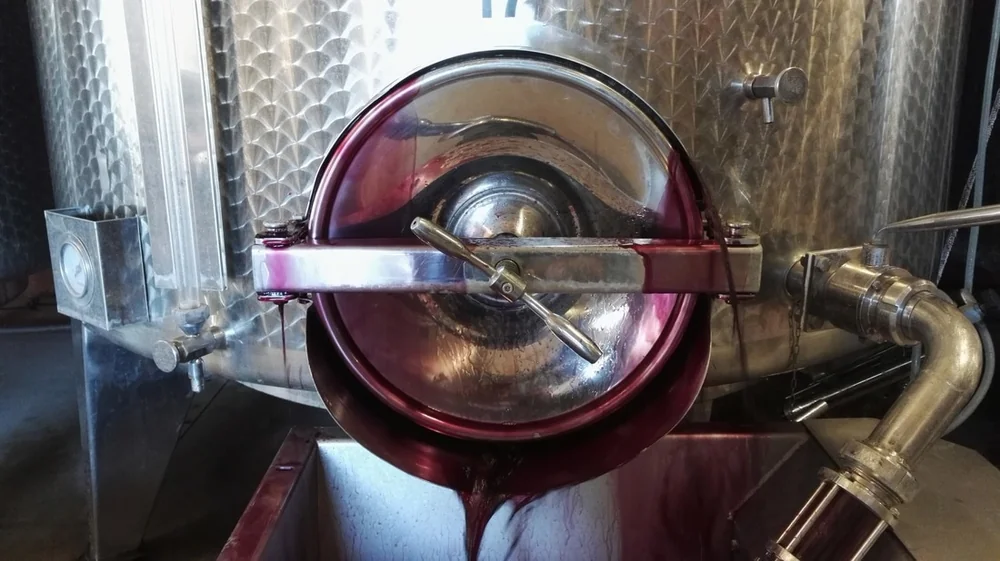 Gros plan d'un vin rouge s'ecoulant d'une vanne de cuve inox vers un bac, eclats de reflets, robe pourpre intense. Image de vinification dynamique et coloree.