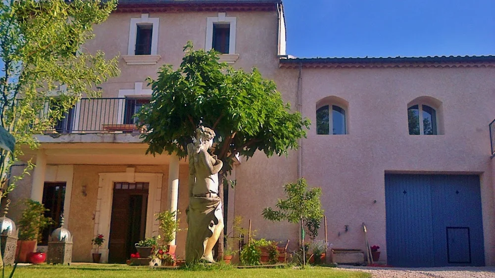 Facade de la maison vigneronne du domaine (ancienne maison de 1824) avec statue, figuier, terrasse a colonnes et grand portail bleu. Belle vue d'ensemble du batiment sous ciel bleu.