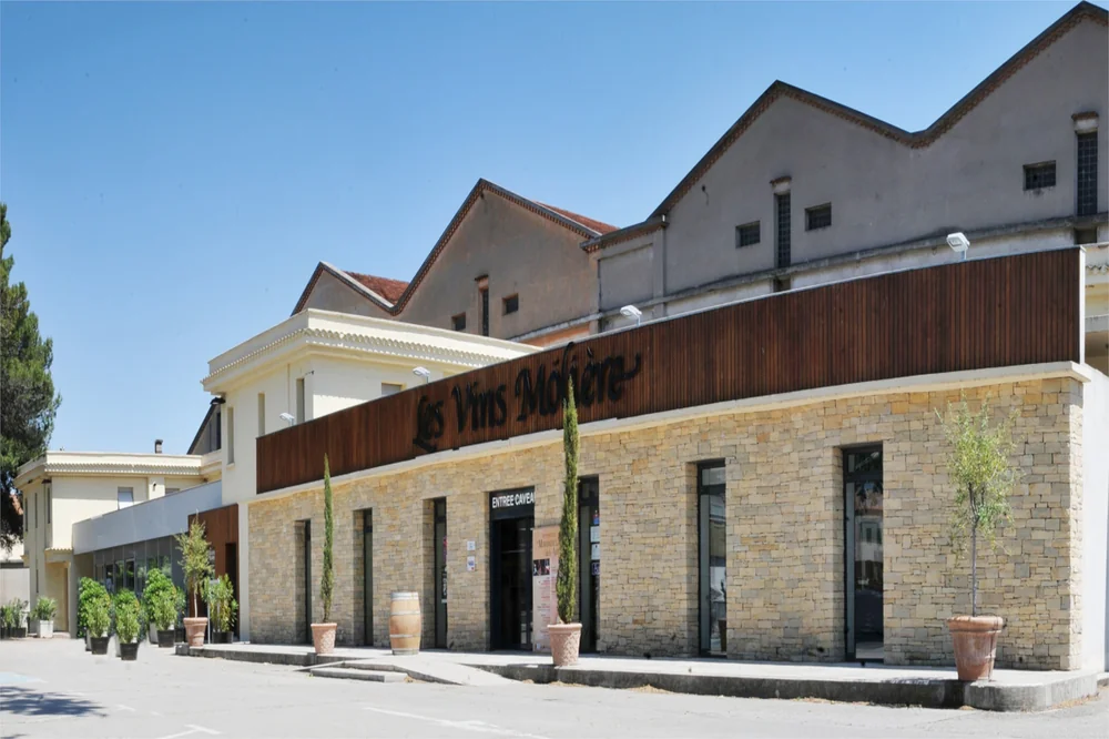 Caveau de Pezenas : facade en pierre avec bardage bois et enseigne 'Les Vins Moliere', alignement de cypres et jarres, ciel bleu. Belle vitrine architecturale du point de vente.