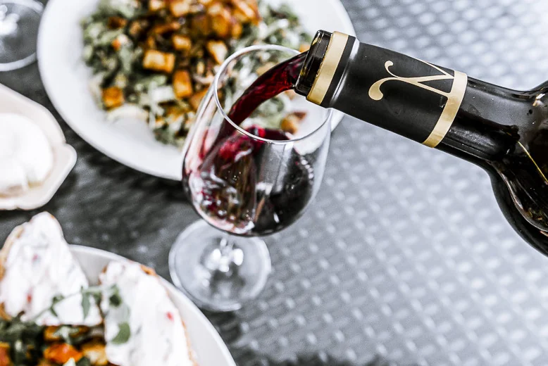 Packshot lifestyle : service d'un vin rouge dans un verre depuis une bouteille (capsule monogramme), entoure d'assiettes de tapas/cuisine. Ambiance art de vivre, accord mets-vin.