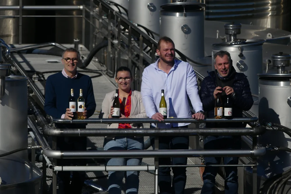 Portrait de groupe de l'equipe / vignerons (4 personnes) posant avec des bouteilles devant les cuves inox du chai, ensoleille. Tres valorisant : met un visage humain sur la cooperative. og:image de la page La Cave.