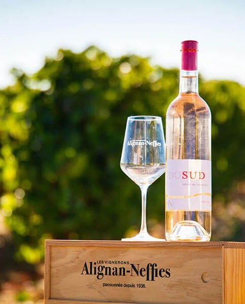 Packshot lifestyle : bouteille de rose 'Du Sud' et verre grave 'Alignan-Neffies' poses sur une caisse bois marquee 'Les Vignerons Alignan-Neffies passionnes depuis 1936', bokeh de vignes. Tres belle mise en situation de marque.