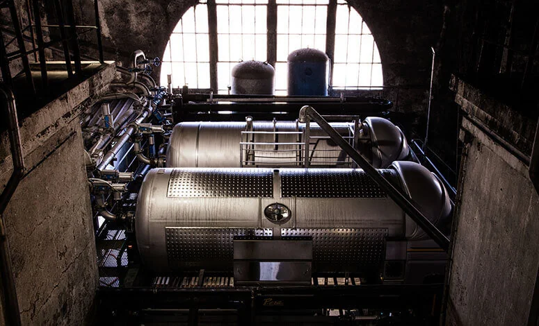 Cuves de vinification en inox alignees sous une grande baie vitree en arc, eclairage atmospherique du chai. Belle image industrielle/technique du chai.