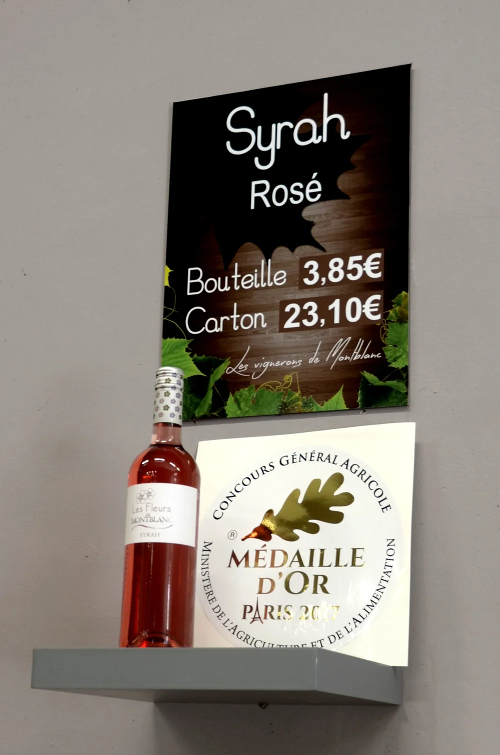 Mise en situation en boutique : pancarte promo 'Syrah Rosé' (Les vignerons de Montblanc), une bouteille 'Les Fleurs de Montblanc' Syrah rose, et a cote l'affichette de la distinction 'Concours General Agricole - Medaille d'Or Paris 2017'. Montre a la fois le produit et une recompense. Photo officiel