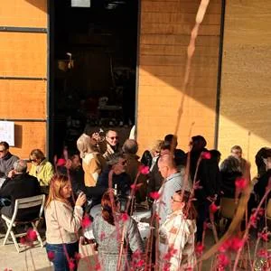 Evenement apero au chai : groupe de visiteurs devant la porte ouverte du chai (bardage bois), branches de fleurs au premier plan. Petite resolution, ambiance plutot que cadrage soigne, mais montre le caveau anime.