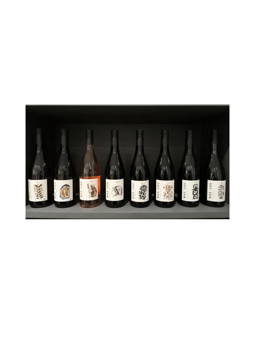 La gamme complete Mas Lou : 8 bouteilles alignees sur une etagere sombre montrant les etiquettes artistiques (rouges, blanc, rose). Packshot d'ensemble en situation, pas une etiquette plate isolee. 1414x2000.