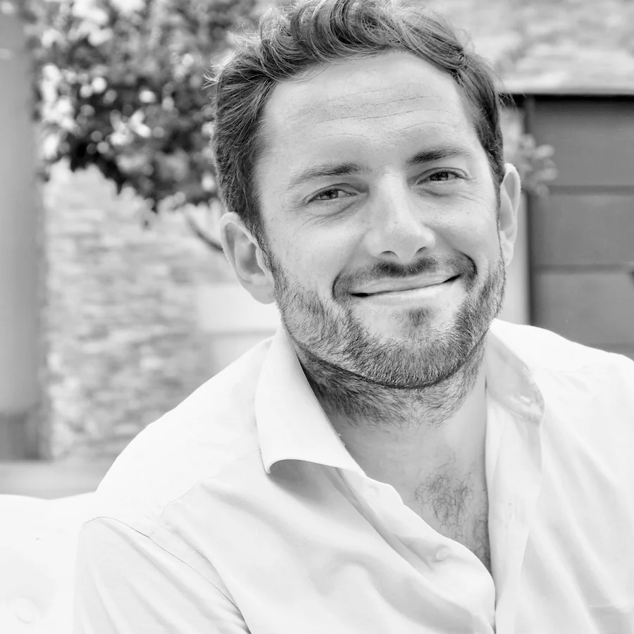Portrait noir et blanc de Romain Frayssinet (gerant du domaine depuis 2010), souriant, chemise blanche, devant un mur de pierre. Tres bon portrait de l'homme cle du domaine (900x900).